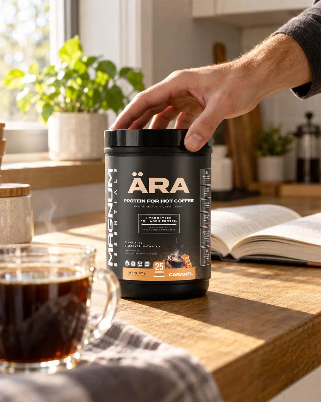A hand reaching for a tub of ÄRA Caramel on a sunny kitchen counter next to a glass of coffee and an open book