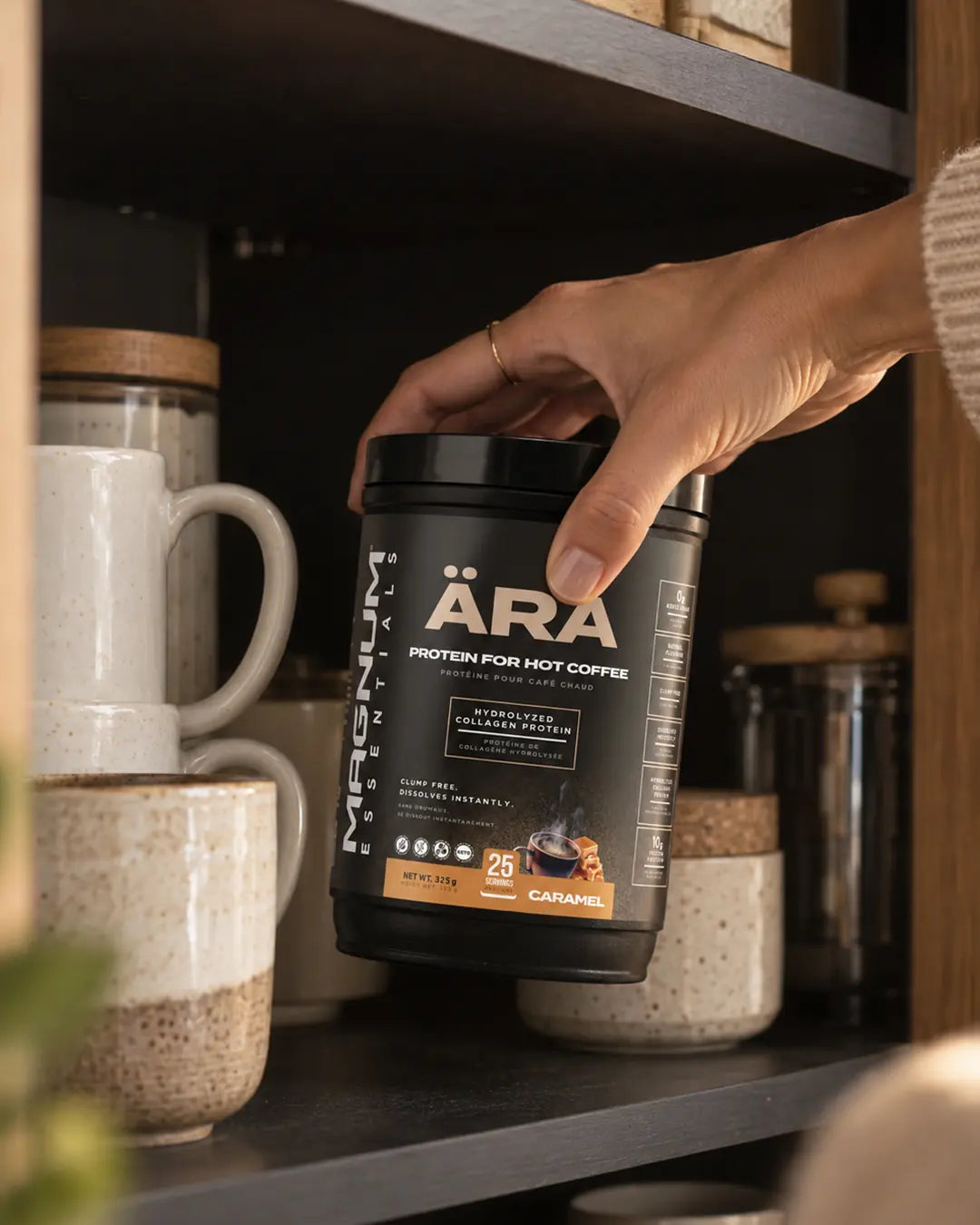 A hand placing a tub of ÄRA Caramel on a kitchen shelf among ceramic mugs and a French press