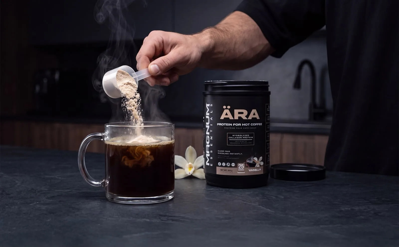 A hand pouring a scoop of ÄRA vanilla powder into a steaming glass mug of black coffee, the ÄRA tub beside it on a dark stone counter